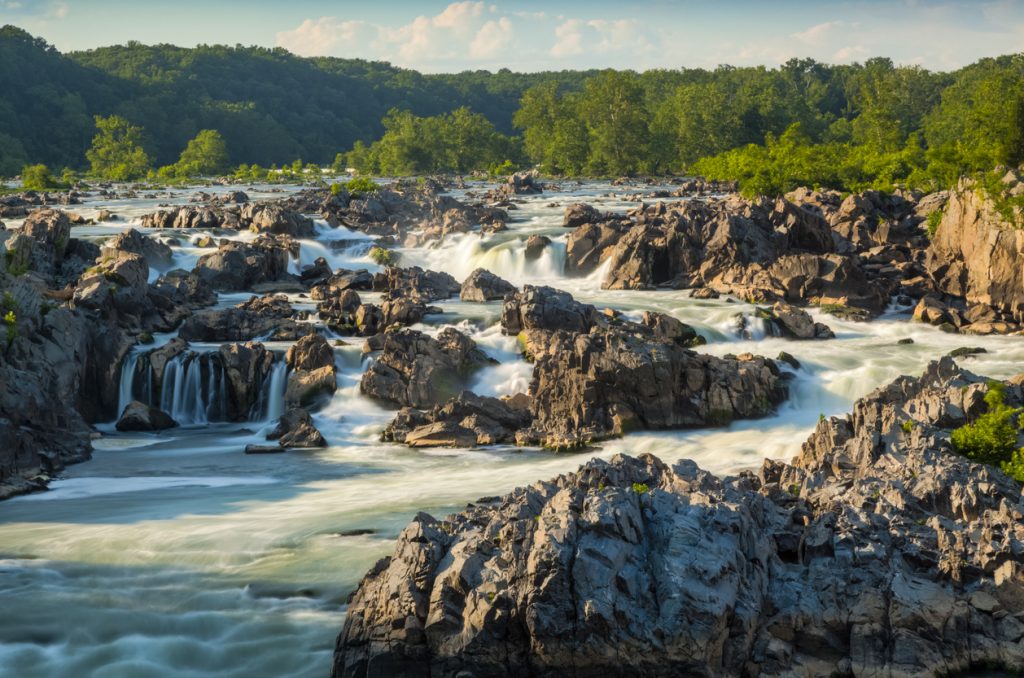 A scenic view of the Great Falls of the Potomac showcases a breathtaking waterfall cascading over rugged rocks, surrounded by lush green trees under a clear blue Maryland sky. The water flows smoothly over the jagged terrain, crafting a serene and natural landscape.