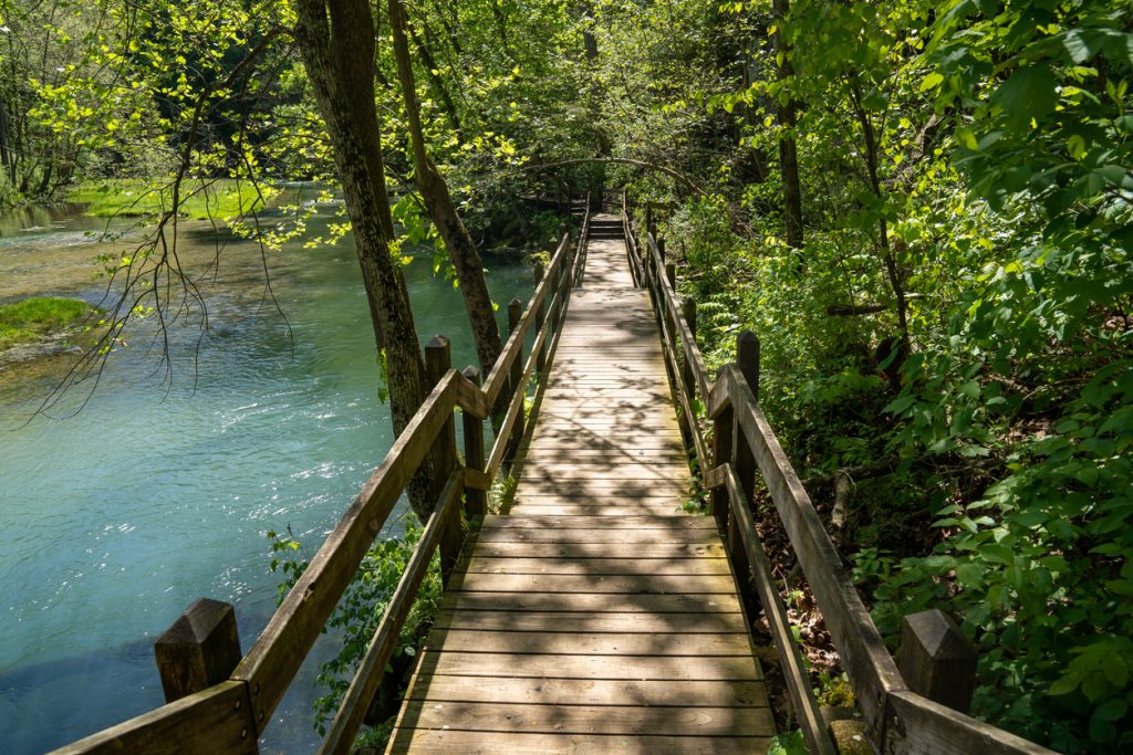 A wooden footbridge crosses over a tranquil stream surrounded by lush green foliage in Ha Ha Tonka State Park, Missouri. Sunlight filters through the trees, casting dappled shadows on the bridge, embodying a peaceful, natural environment.