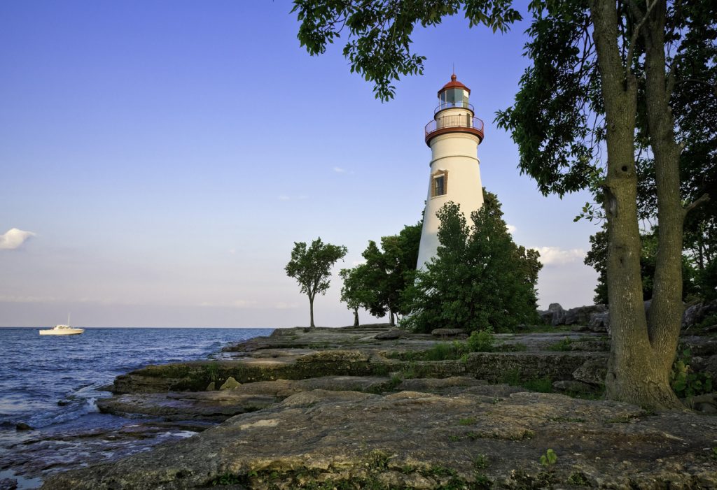 The Marblehead Lighthouse, with its distinctive white structure and red top, majestically stands on Ohio's rocky Lake Erie shoreline, surrounded by lush green trees. A small boat peacefully drifts on the calm blue water under a clear sky.
