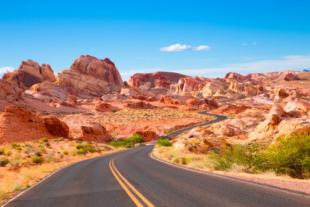 A winding road stretches through Nevada's vibrant desert landscape in the Valley of Fire, with red and orange rock formations under a clear blue sky. Sparse green shrubs dot the area, creating a scenic view of a sunny, arid terrain.