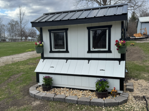 A small, white backyard chicken coop with black trim, two windows, and hanging flower pots. The coop sits on a border of rocks and bricks, surrounded by potted flowers, with grass and cloudy skies in the background.