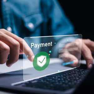 A person uses a laptop with a virtual payment interface displayed; a hand points to a floating screen showing the word "Payment" and a green shield with a checkmark, suggesting secure online payment.