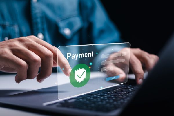A person uses a laptop with a virtual payment interface displayed; a hand points to a floating screen showing the word "Payment" and a green shield with a checkmark, suggesting secure online payment.