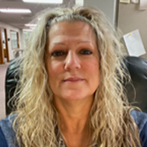 RoseAnn with long, curly blonde hair is sitting in an office chair, looking at the camera. The background shows an office hallway and some papers pinned to a wall.
