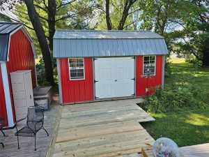 A red shed with a gray metal roof and white double doors sits beside a wooden deck, surrounded by green grass, trees, and outdoor furniture on a sunny day.